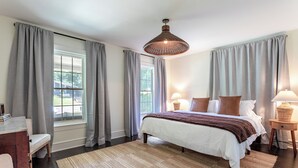 Un dormitorio con una cama grande, dos mesitas de noche con lámparas, un ventanal con cortinas y suelo de madera.