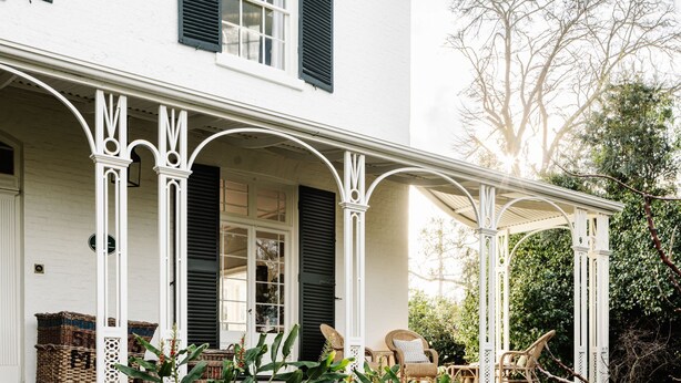 A white building with black shutters, a covered porch with wicker furniture, and a garden with trees and plants.