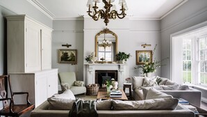 A spacious living room with a fireplace, a large mirror, and a chandelier. There are multiple seating areas and a view of the outdoors through large windows.