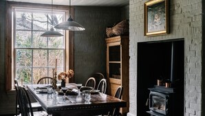 A dining room with a wooden table, chairs, hanging pendant lights, a fireplace, and a large window with a view of trees.