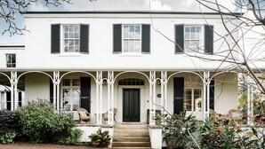 A two-story white building with black shutters, a central black door, and a porch with white columns and wicker furniture.