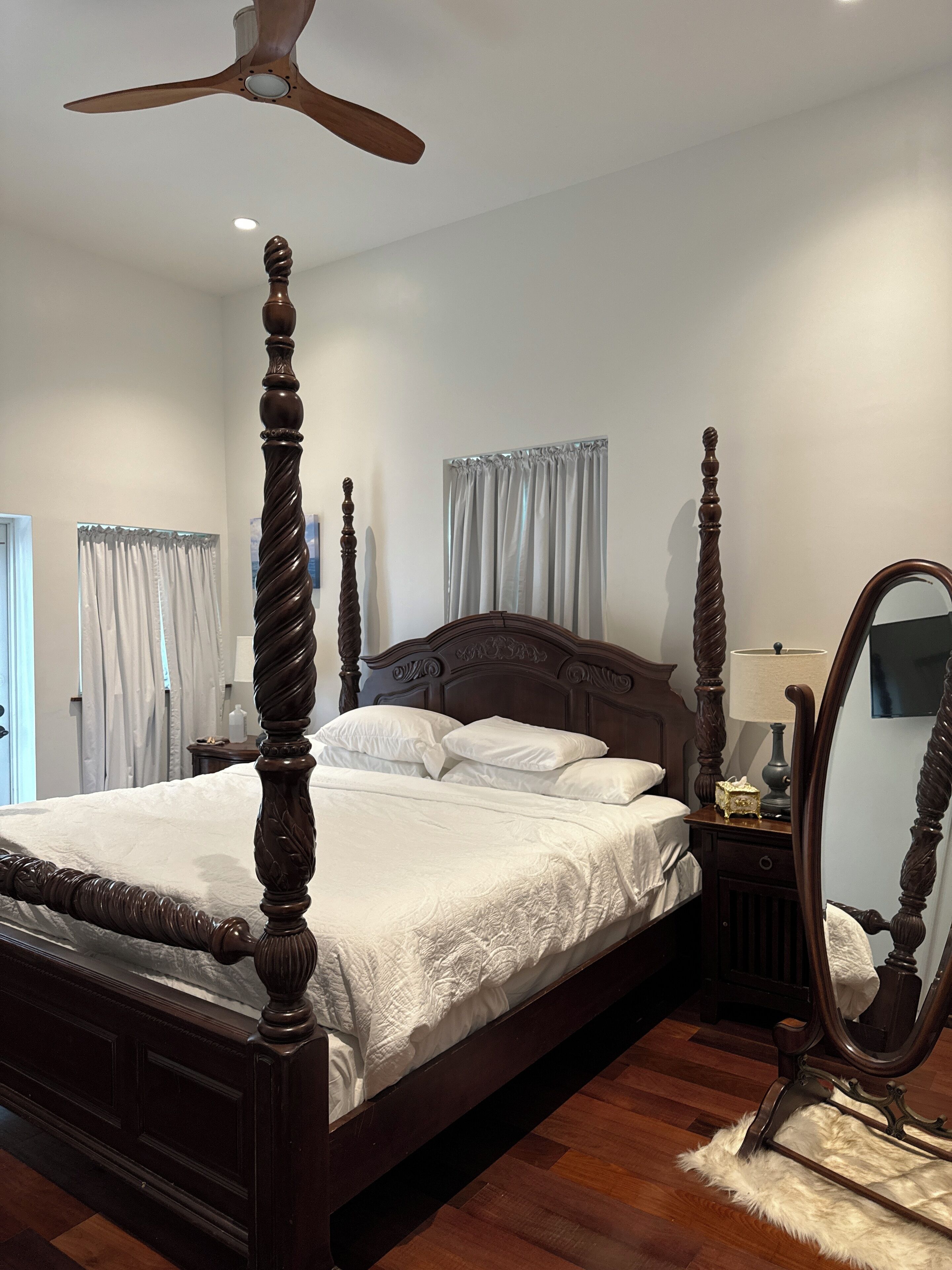 A four-poster bed with a wooden headboard and footboard, a ceiling fan, a bedside table with a lamp, and a mirror.