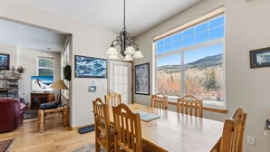 Un comedor amplio con una gran mesa de madera y sillas, una araña y ventanas grandes que ofrecen vistas a un paisaje montañoso.