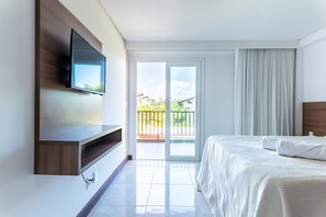 Habitación de hotel moderna con una cama grande, un televisor de pantalla plana montado en un panel de madera y una puerta de cristal corredera que da a un balcón con vistas a zonas verdes.