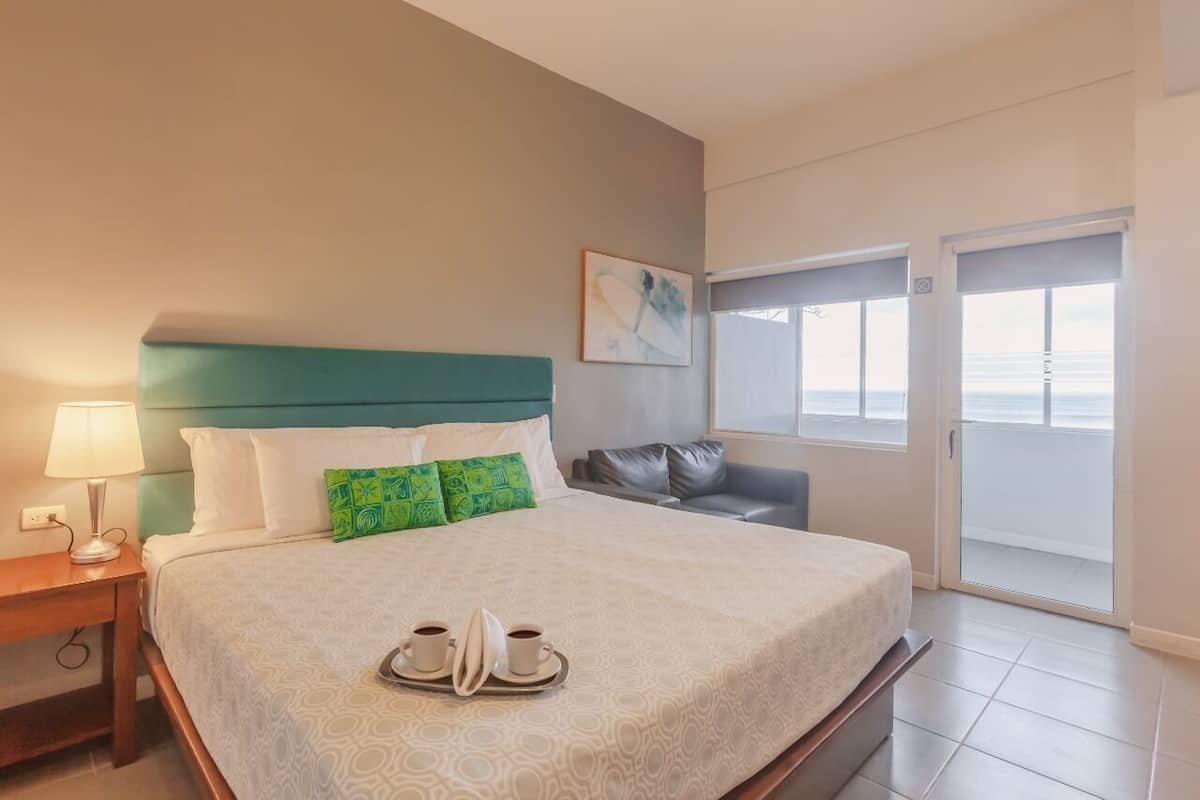 A hotel room with a bed, a nightstand, a lamp, a tray with cups, a green pillow, a gray sofa, and a view of the ocean through a sliding door.