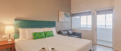 Une chambre d’hôtel avec un lit, une table de chevet, une lampe, un plateau avec des tasses, un oreiller vert, un canapé gris et une vue sur l’océan par une porte coulissante.