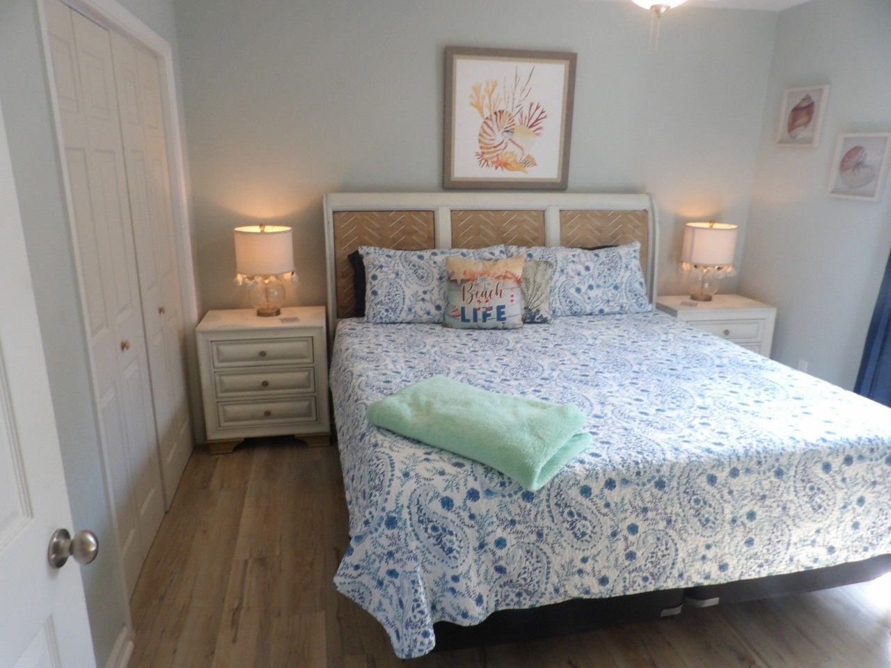 Quarto com cama, duas mesas de cabeceira, um criado-mudo, uma porta e um quadro na parede.