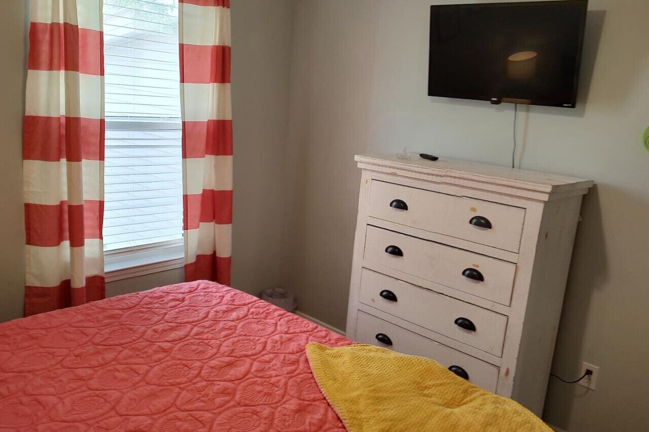 A bedroom with a bed, a dresser, a television on the wall, and a window with curtains.