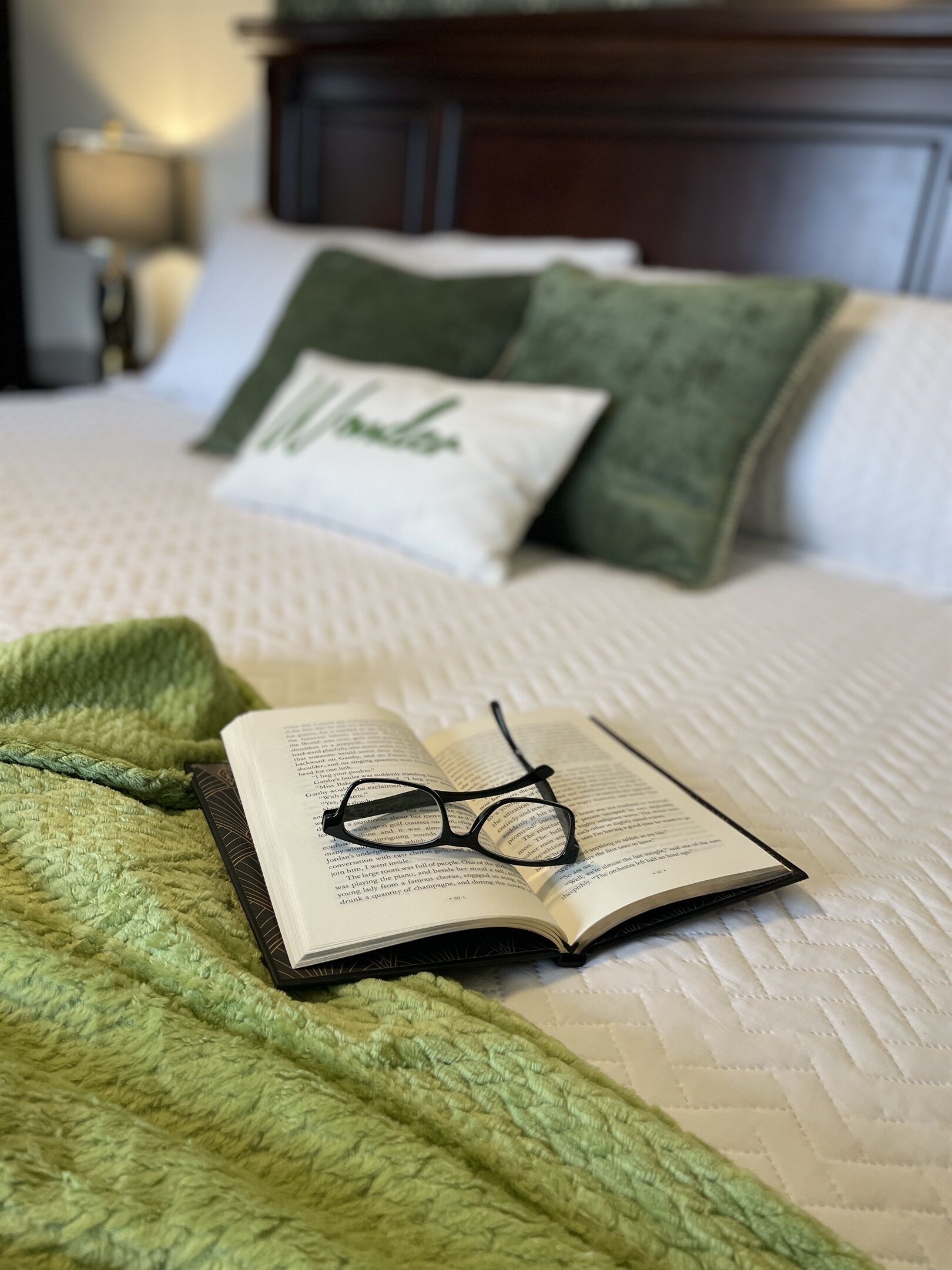 Un lit avec une couverture verte, un oreiller sur lequel est écrit le mot « Wonder », et un livre sur lequel reposent des lunettes.