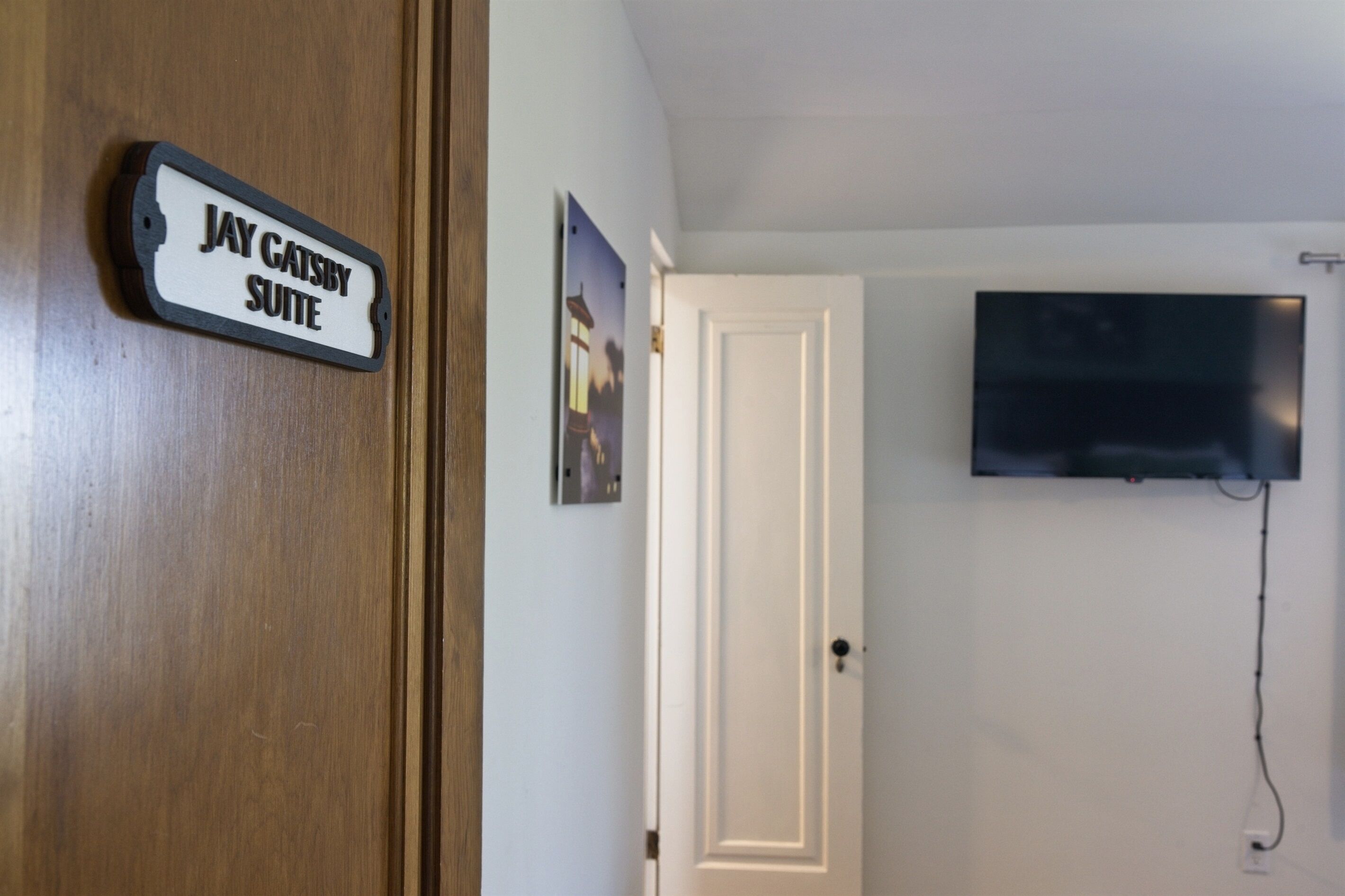 A room with a sign reading "Jay Gatsby Suite," a mounted TV, and a door.