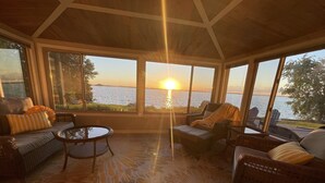 A sunlit room with a view of the sunset over water, furnished with wicker sofas and chairs, and a patterned carpet.