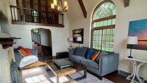 A spacious living room with a high ceiling, a staircase leading to an upper level, a large window, and a painting on the wall.
