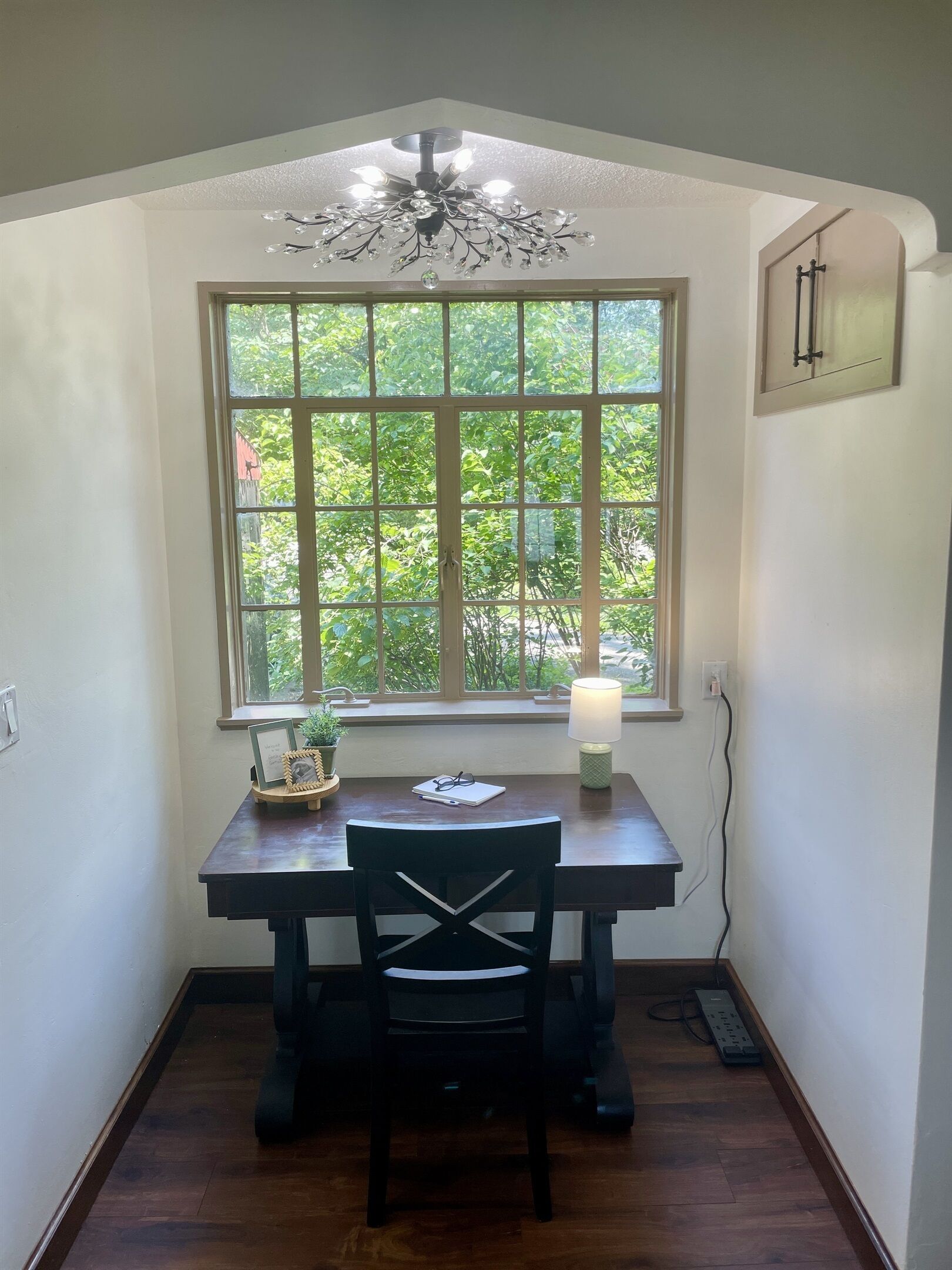 A corner room with a wooden desk, a black chair, a window with a view of greenery, a chandelier, and a wall clock.