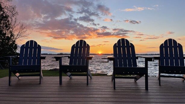 Three Adirondack chairs on a wooden deck with a view of the sunset over a body of water.