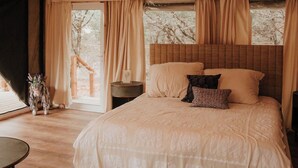 A spacious bedroom with a large bed, a wooden floor, a ceiling light fixture, and a view of trees through the window. - Lavender - Luxury Glamping Tent (Johnson City)