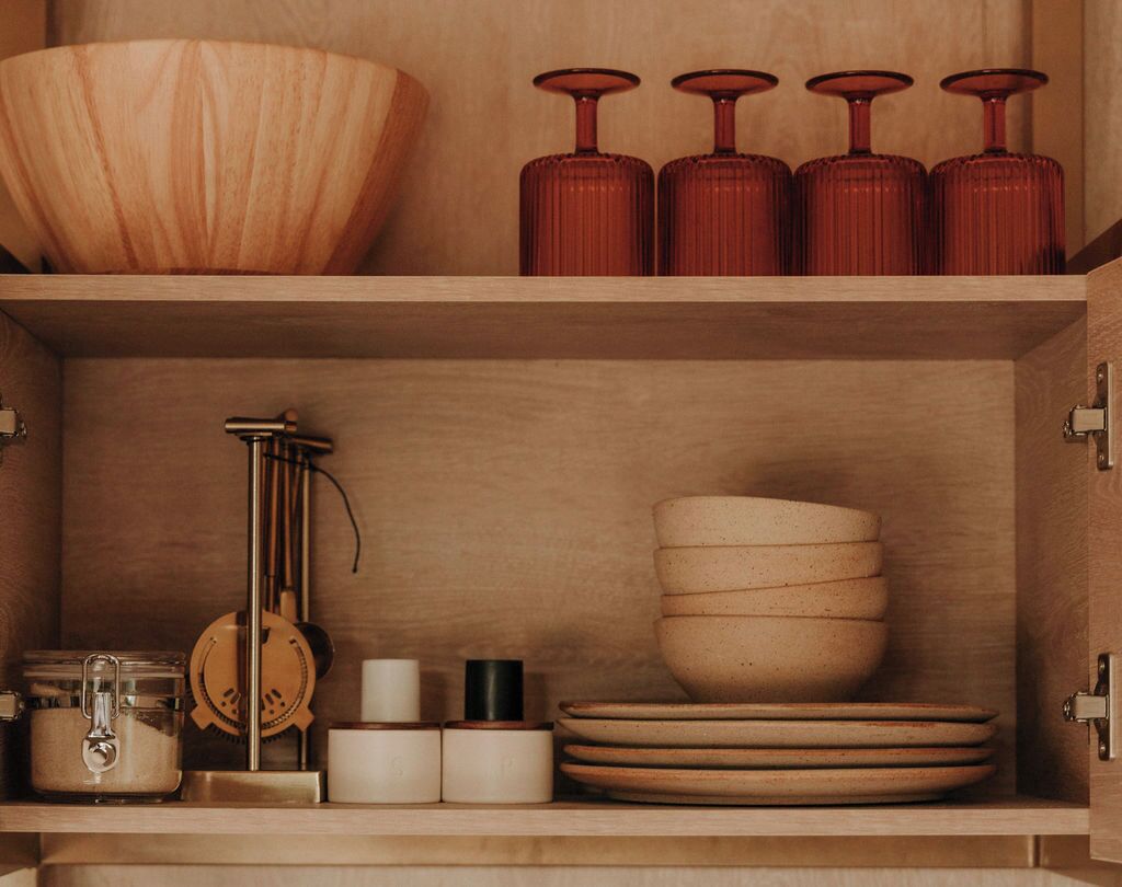 A wooden cabinet with shelves containing kitchenware, including a large bowl, several plates, and red glass bottles.