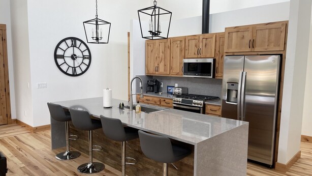 Cozinha moderna com ilha central, banquetas, armários de madeira, eletrodomésticos de aço inoxidável e iluminação pendente.
