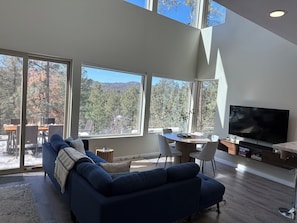 Un salon moderne doté d’un grand téléviseur, d’un canapé et d’un coin repas offrant une vue sur les arbres et les montagnes.