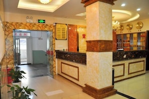 Un hall de hotel con un mostrador de recepción, una entrada con puerta de vidrio y un área de recepción con una columna de mármol.