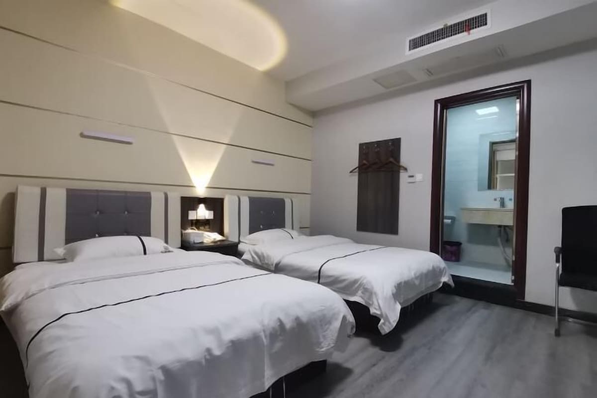 A hotel room with two beds, a bedside table with a lamp, a mirror, and a bathroom visible through an open door.