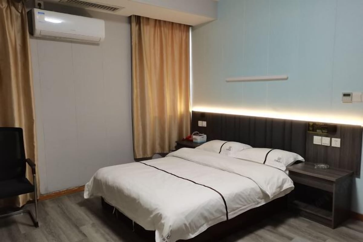 A hotel room with a bed, a chair, a nightstand, an air conditioner, and a wall-mounted shelf.