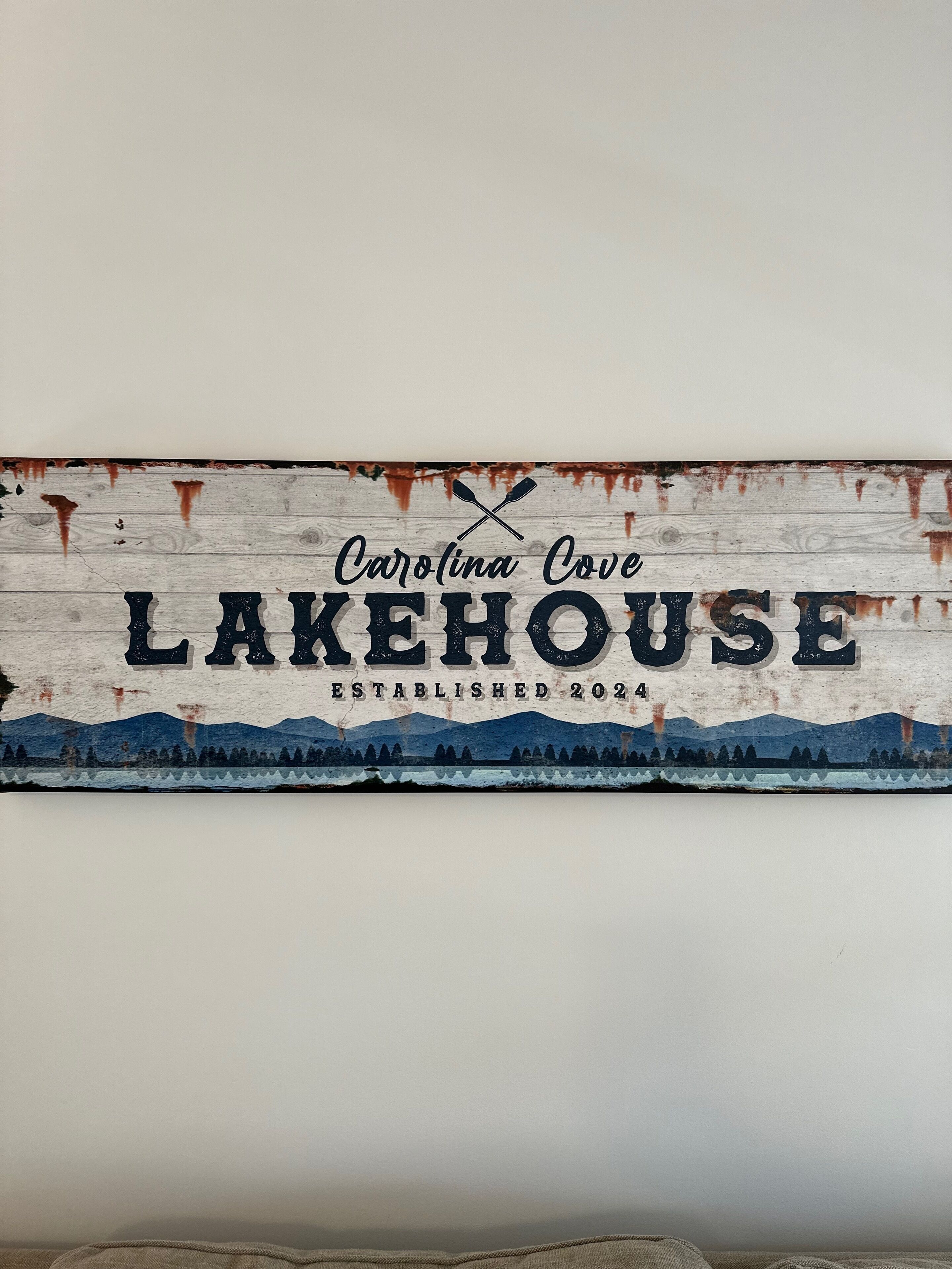 En rustik skylt med texten ”Carolina Cove Lakehouse, grundat 2024” och en illustration av ett berglandskap.
