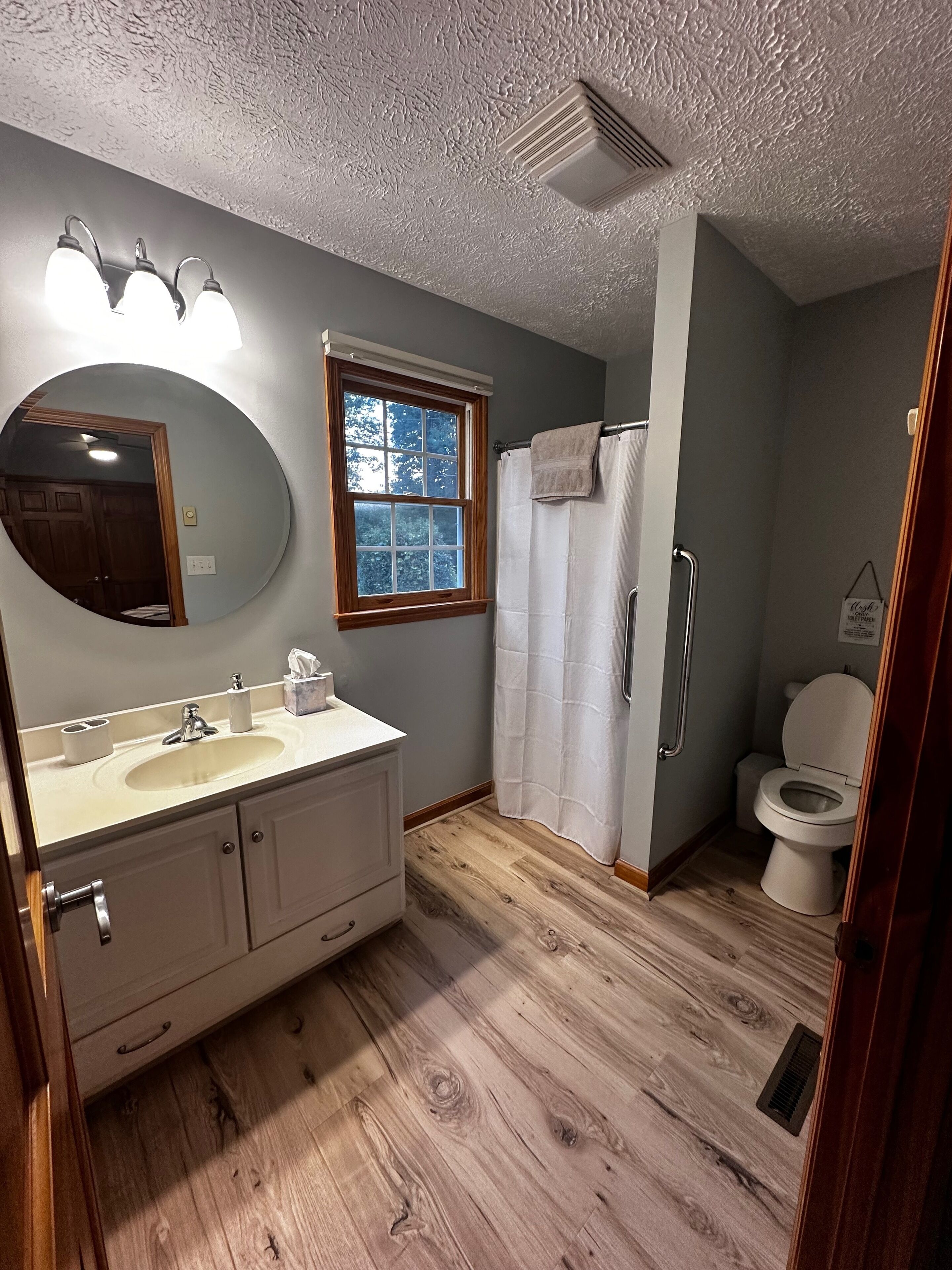 A bathroom with a round mirror, a sink, a window, a shower curtain, and a toilet.