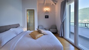 Un dormitorio con una cama grande, una puerta con un panel de vidrio emplumado, una lámpara fijada a la pared y un balcón con vistas al paisaje.