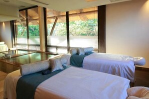 Habitación de hotel con dos camas, una camilla de masaje y ventanas amplias con persianas.