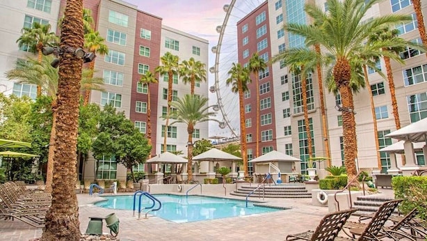 A hotel pool area with palm trees, lounge chairs, and a building with multiple floors and windows.