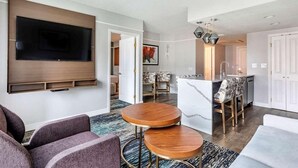 Un salon moderne doté d’un téléviseur à écran plat fixé au mur, d’un coin salon avec un canapé et deux petites tables rondes en bois, et d’un espace cuisine avec un comptoir en marbre et des tabourets de bar.