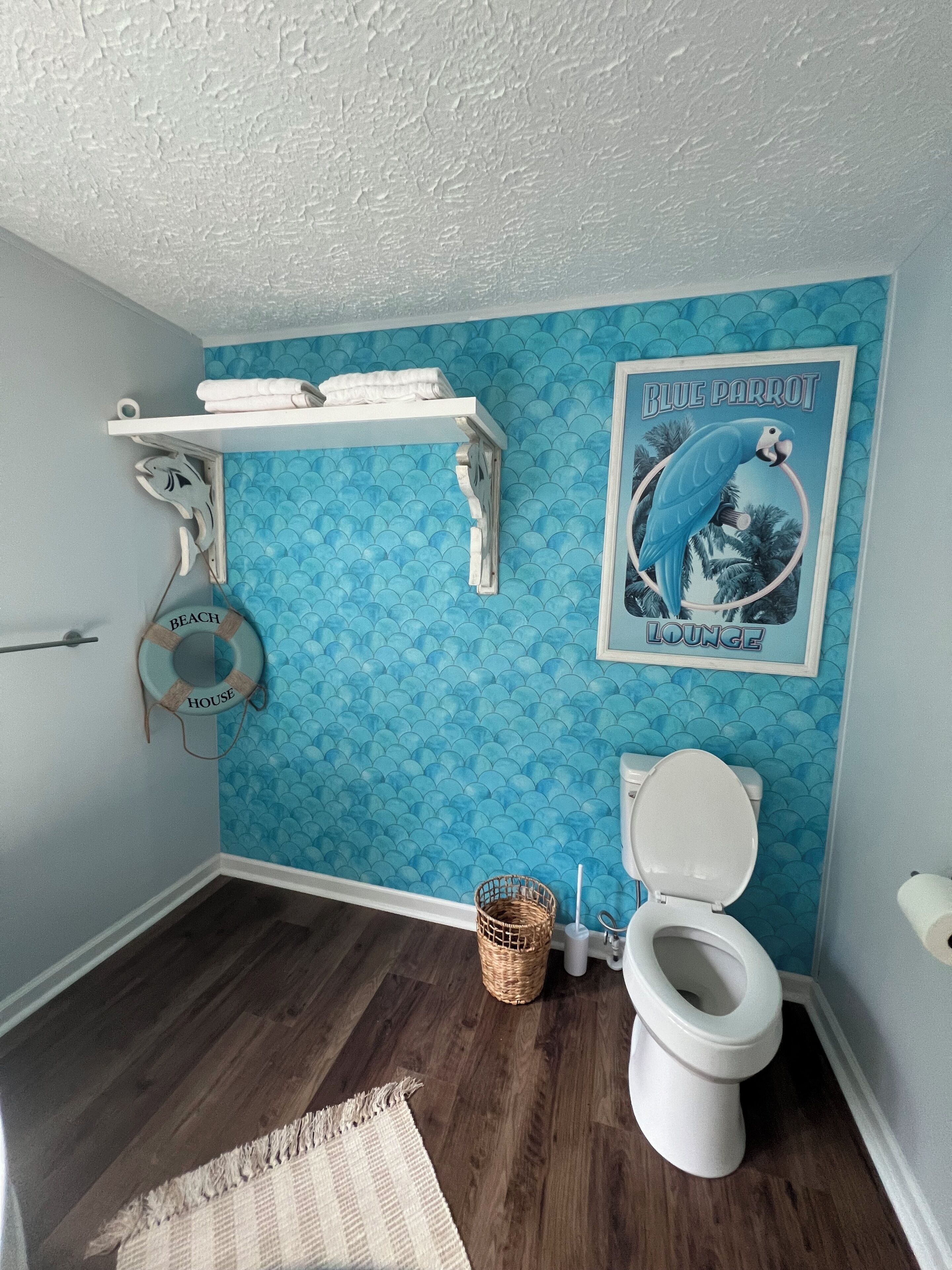 A bathroom with a blue tiled wall, a white toilet, a white shelf with towels, a life preserver decoration, and a poster of a parrot.