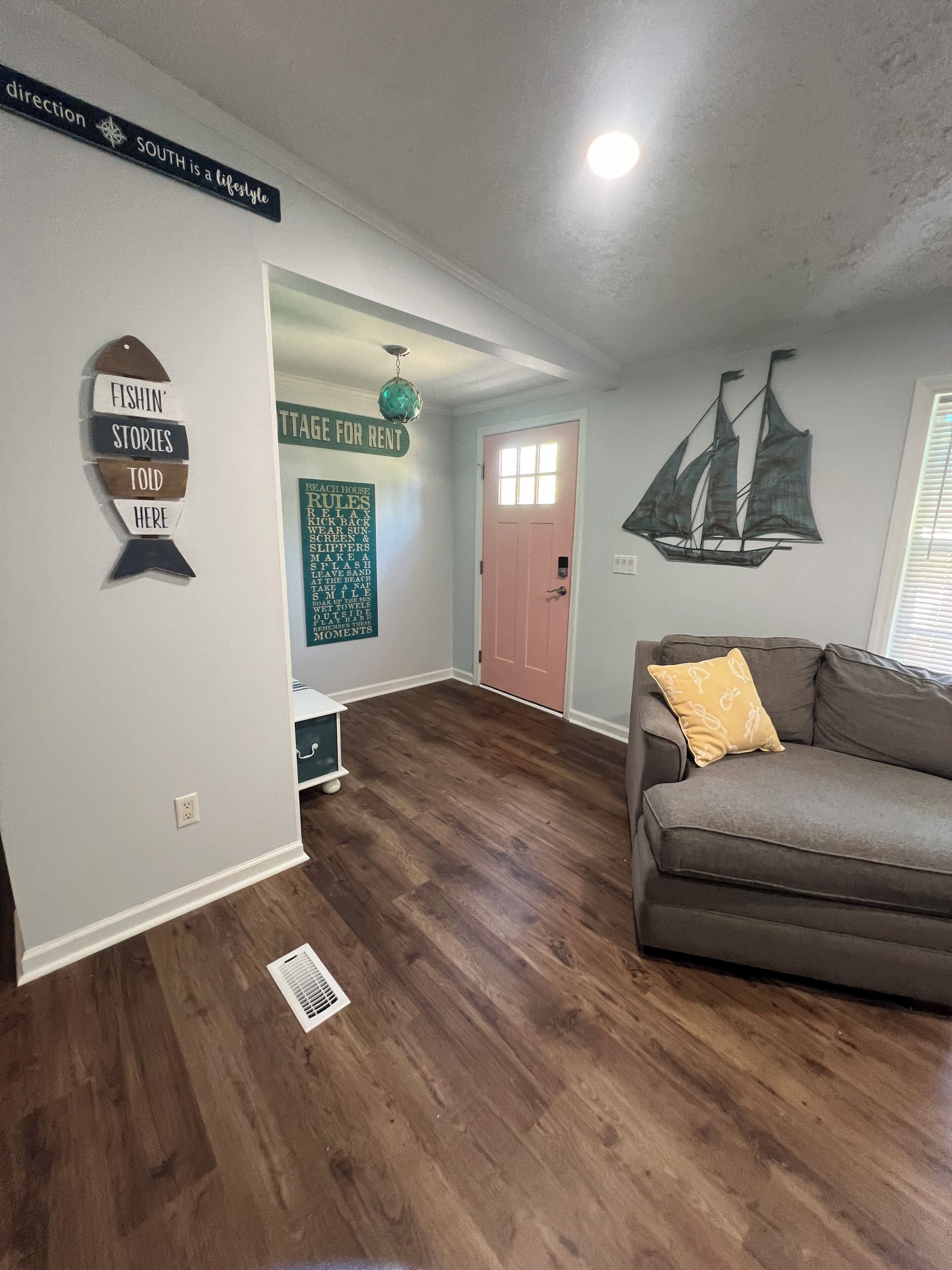 Una sala de estar con piso de madera, un sofá gris con un cojín amarillo, una decoración náutica en la pared y un letrero que dice "direction SOUTH is a lifestyle".