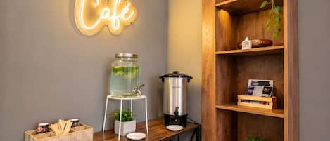 Un café aménagé avec un panneau néon, un distributeur d’eau, une machine à café et une étagère en bois où sont posés des livres et une plante.
