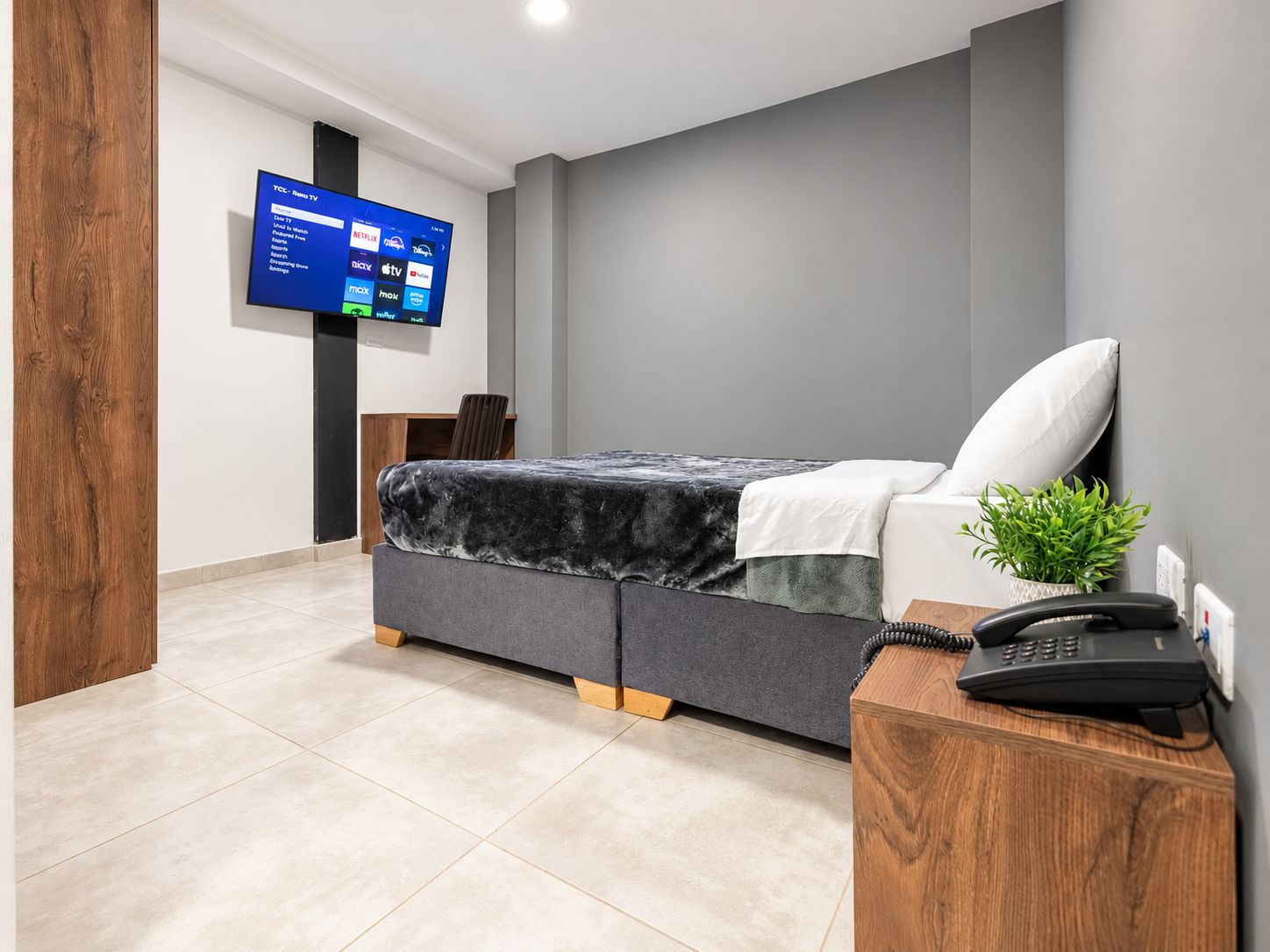 Un dormitorio moderno con una cama, una mesita de noche con un teléfono, un televisor montado en la pared y una pequeña planta.