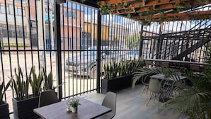 Ein möblierter Patio-Bereich im Freien, mit Holztisch und -stühlen, umgeben von Blumentöpfen und einem Metallzaun.