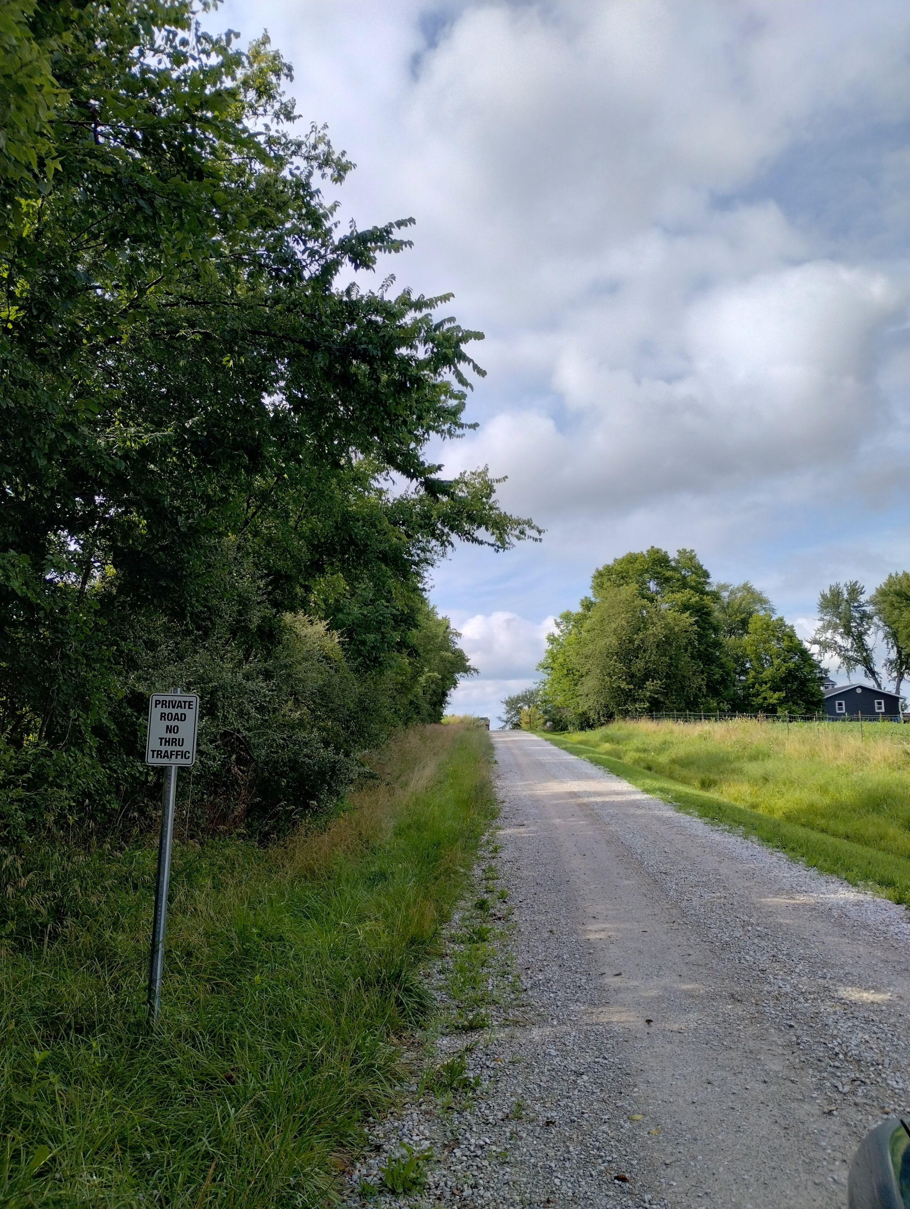 Un camino de grava con un cartel que dice 'PRIVATE ROAD NO THRU TRAFFIC', rodeado de vegetación y un cielo nublado.