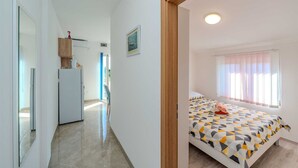 A bedroom with a bed, a window with curtains, a door, and a hallway leading to a kitchen area. - Emila's Apartment- Blue (Nin)