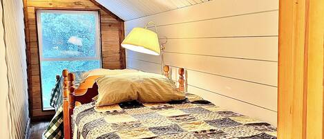 A bedroom with a wooden sloped ceiling, a large window, a bed with a quilt, and a floor lamp.