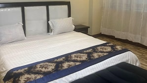 A neatly made bed with a patterned bedspread, a bedside table, and a window with curtains. - ROYAL COMPLEX HOTEL AND APARTMENTS (Kampala)
