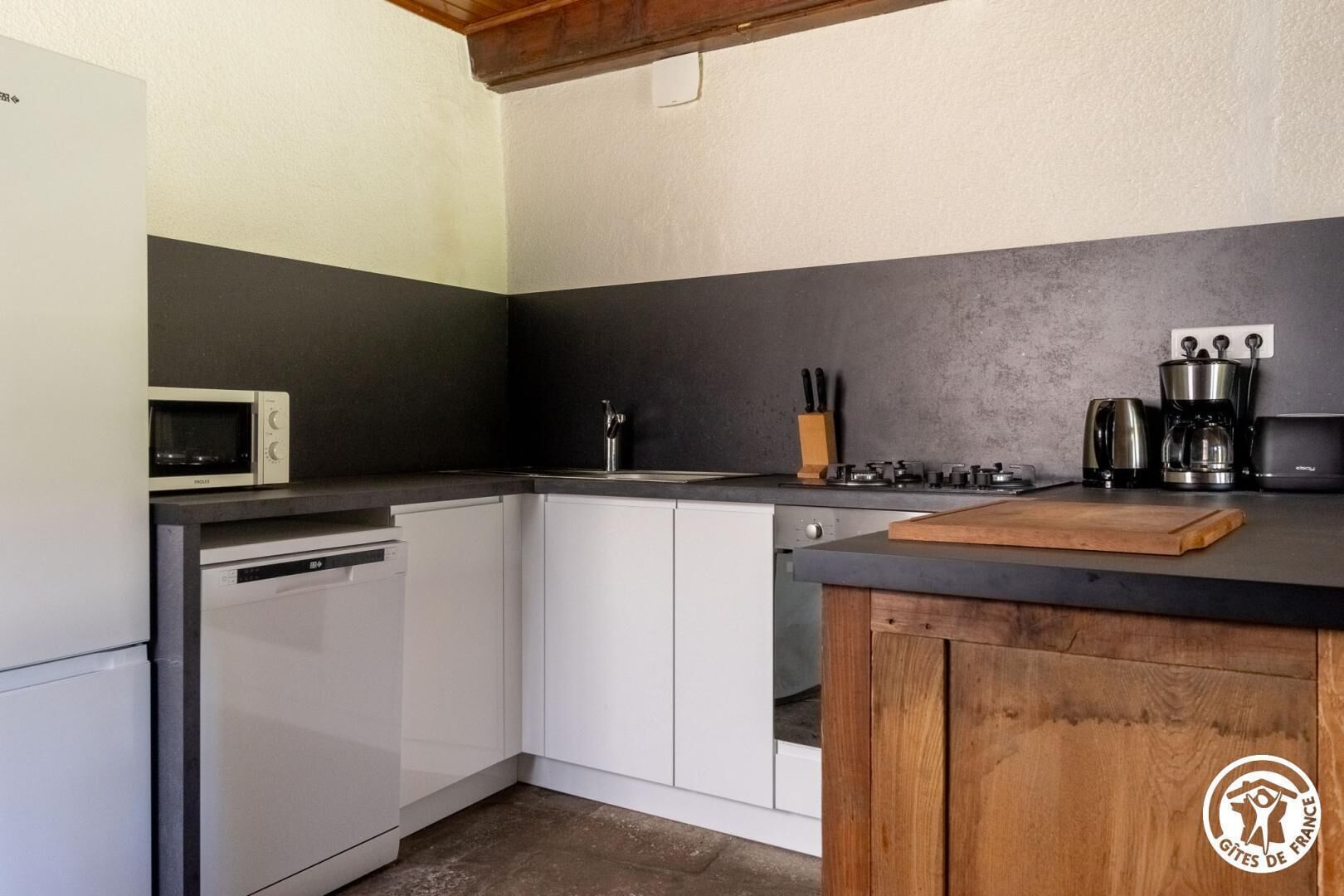 Een kleine keuken met witte kastjes, een magnetron, een koffiezetapparaat en een houten snijplank.