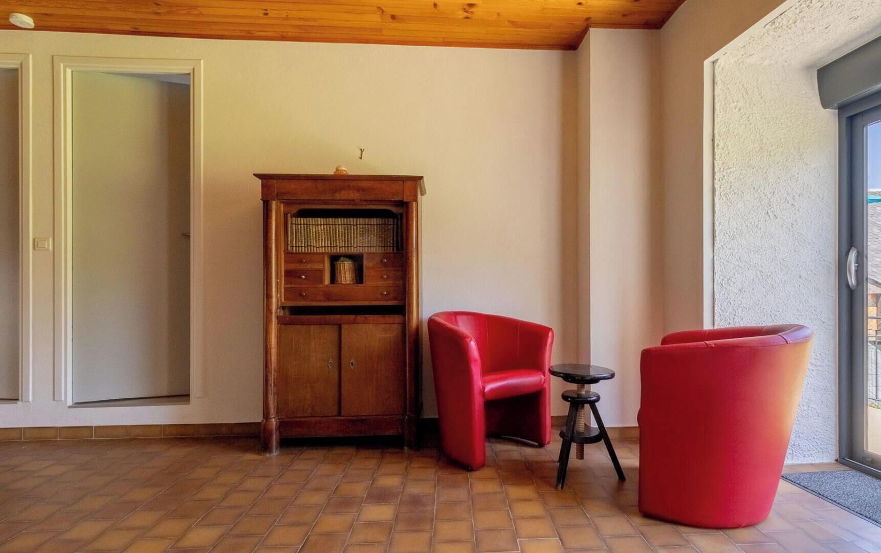 Ein Zimmer mit einem Holzkasten, zwei roten Stühlen, einem kleinen Tisch und einer Tür.