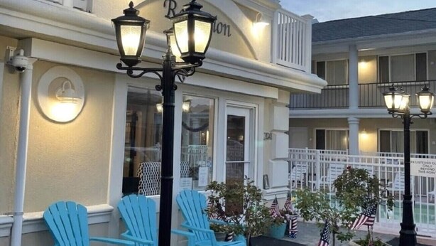 A building with a white facade, blue Adirondack chairs, and American flags in planters. - The Lamplight Inn (Lavallette)