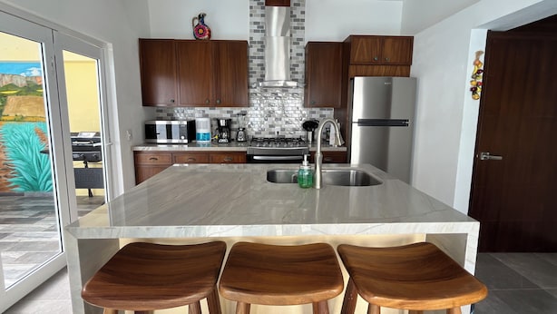 A modern kitchen with a central island, wooden bar stools, stainless steel appliances, and a view of a landscaped area. - Casa Sueños in Bucerias Mexico with Private Pool and Private Parking Garage.  (Bucerías)