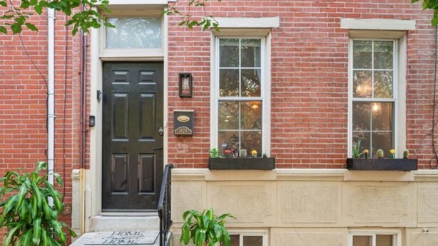 A brick building with a black front door, a small porch with steps, and two windows with flower boxes.