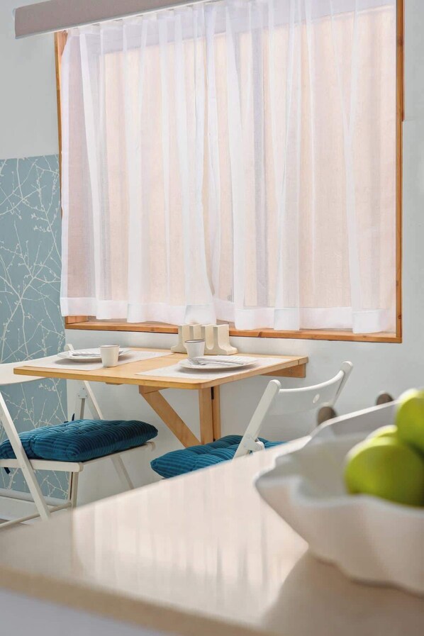 A dining area with a table set for two, a window with sheer curtains, and a view of the outdoors.