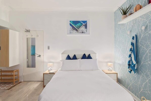 A modern bedroom with a bed, a door, a small table with a lamp, and a wall with a plant and a picture.