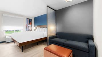 Moderni hotellihuone, jossa on suuri sänky, sohva, pieni jalkarahi ja ikkuna, josta avautuu näkymä parkkipaikalle.