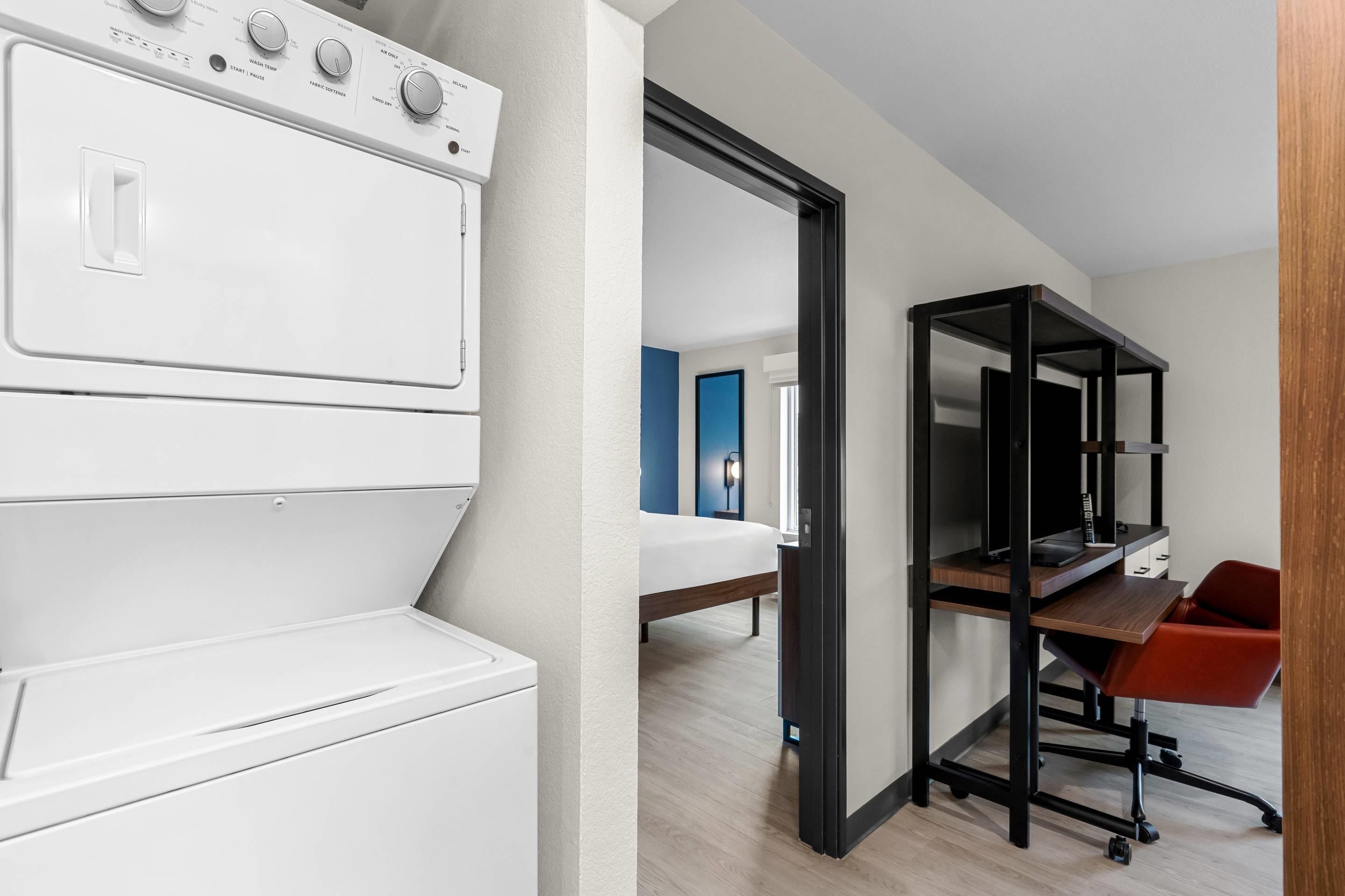 Quarto de hotel moderno com máquina de lavar roupa branca, uma cama, secretária com cadeira e estante preta.