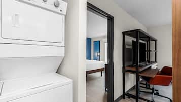 Una habitación de hotel moderna con una lavadora blanca, una cama, un escritorio con una silla y un estante negro.